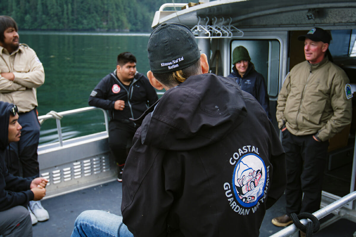 group of six men and women talk in a circle onthe deck of a boat. in the foreground, the back of one person's jacket reads "Coastal Guardian Watchmen"