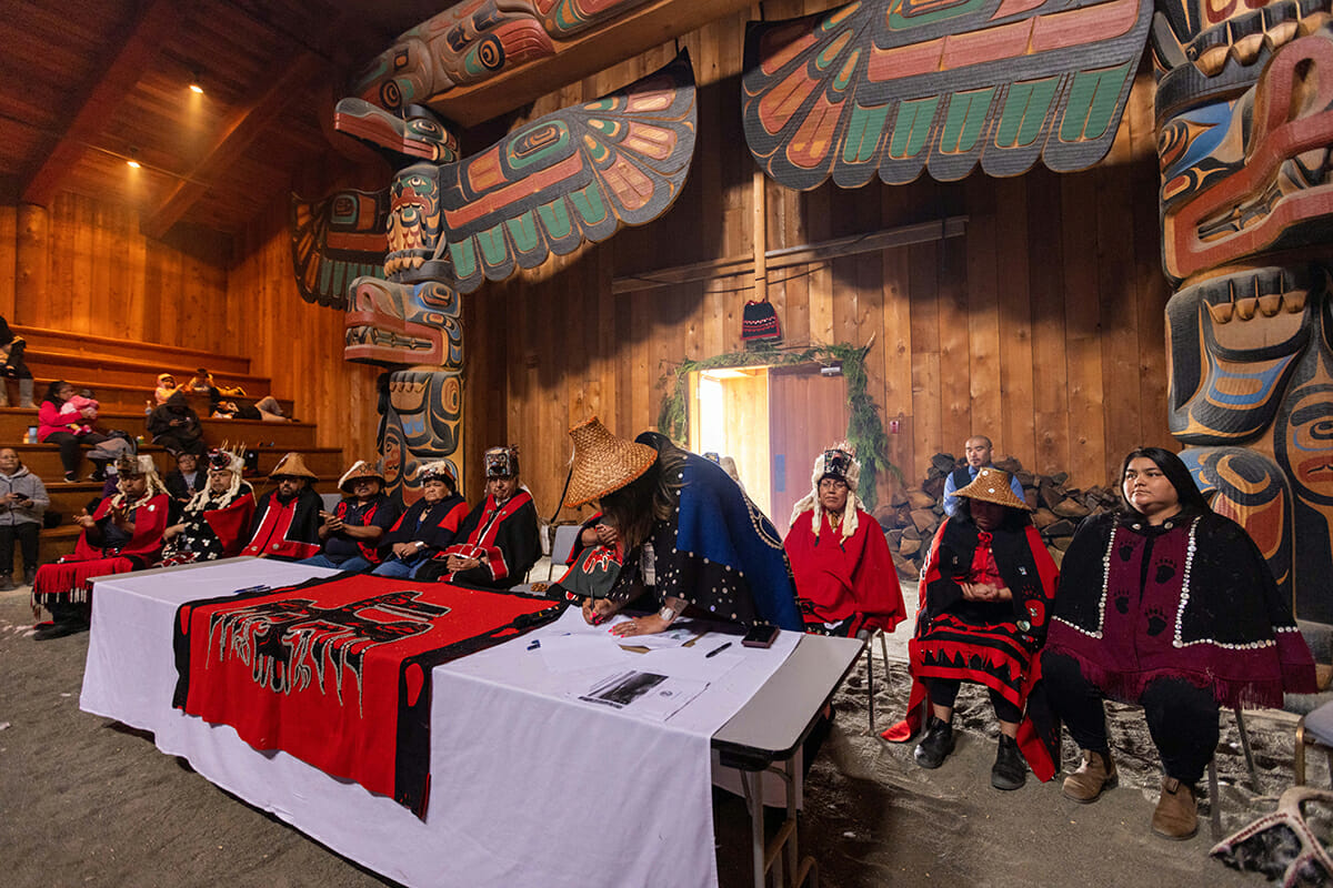 person wearing a cedar hat and regalia leansover a table to sign a declaration. 