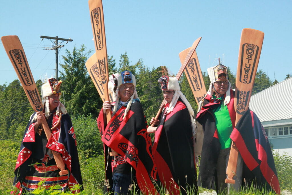 hereditary chiefs in regalia (red and black blankets, white headdresses) carry painted canoe paddles
