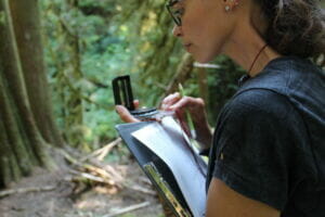 woman faces away from camera and stands in a forest, clipboard in hand