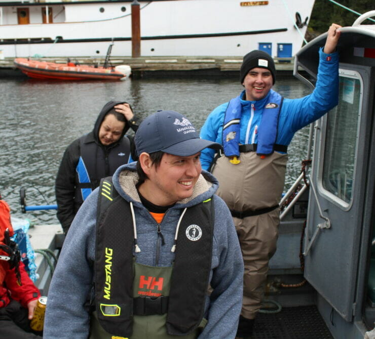 Four people are wearing PFDs and standing on the back of an aluminum boat. The man in front is smiling and wearing a ballcap that says 
