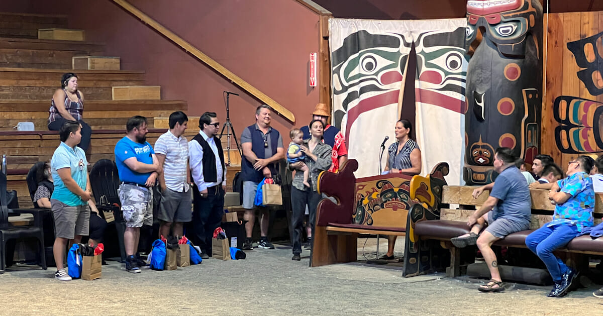 group of young adults stand in a line at the left, while a young woman with a baby on her hip speaks into a microphone. they're in a large wood-beam building decorated with totem poles and formline art
