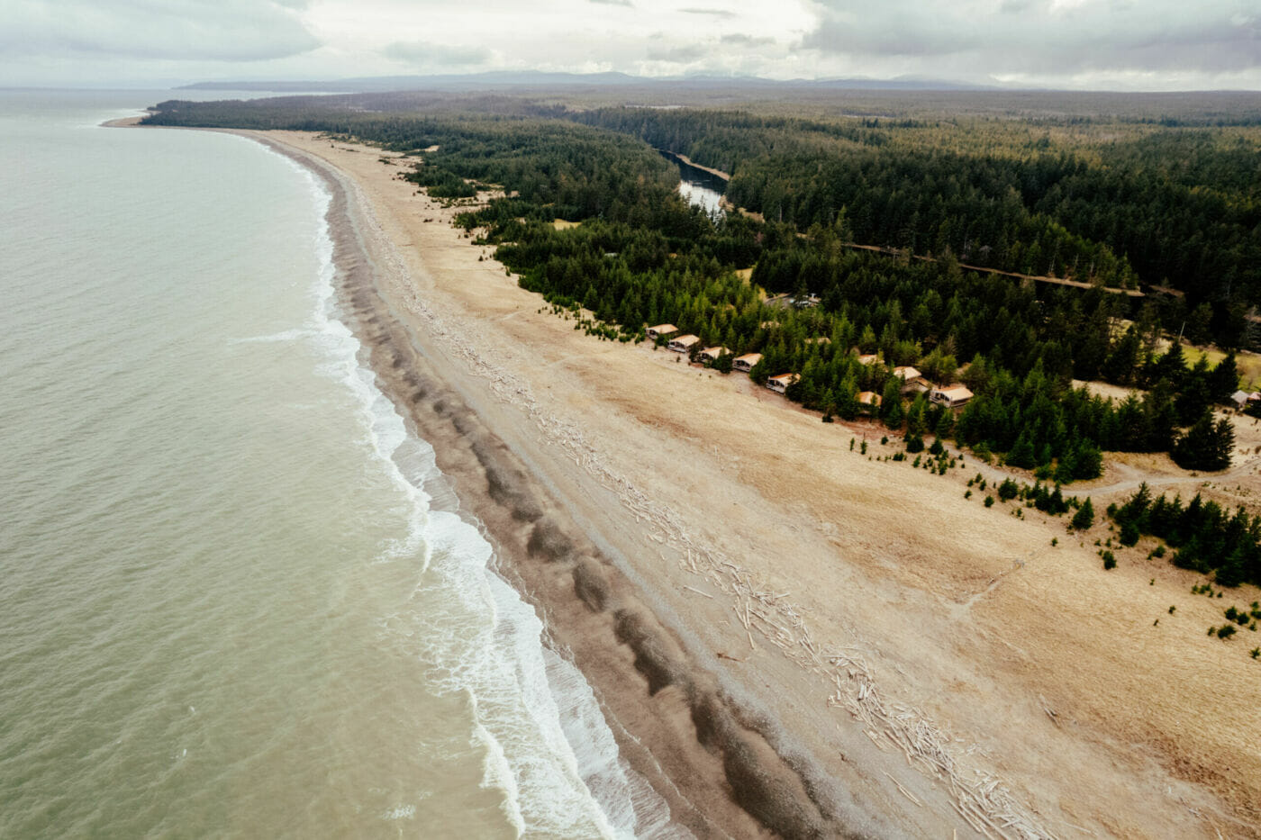 aerial view of a sandy shoreline with small cabins between the woods and the beach
