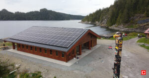 Drone photo of a new building topped with solar panels. In the foreground, a carved and painted pole stands sentry over the shoreline.