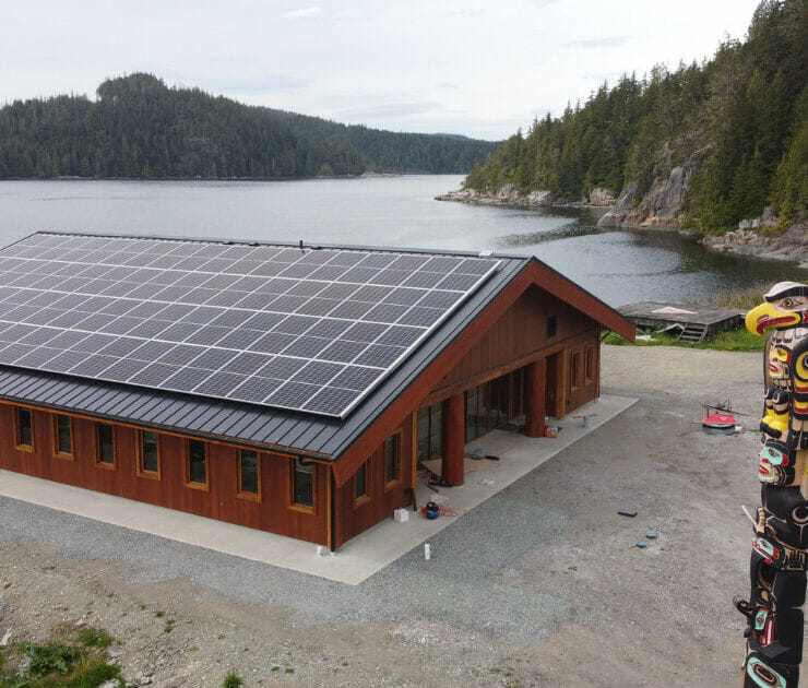 Drone photo of a new building topped with solar panels. In the foreground, a carved and painted pole stands sentry over the shoreline.