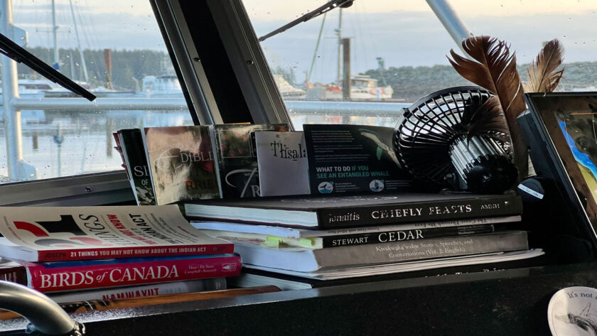a pile of books about Indigenous culture, cedar, and wildlife sit on the dashboard of a boat