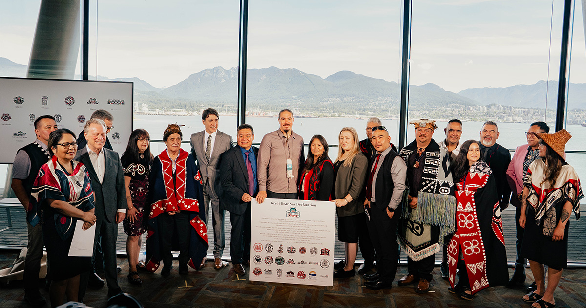 A group of leaders from 17 First Nations stand together around a large plaque. The plaque says "Great Bear Sea Declaration" and below it is a list of statements and logos from the 17 Nations. All the leaders standing together are smiling proudly. Some leaders are wearing regalia, others are wearing business clothing. Joined by the leaders are other officials, including the former Prime Minister Justin Trudeau.