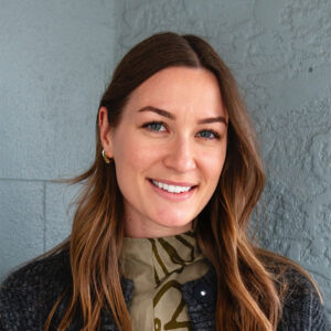 A portrait image of Logan Griffiths. She is smiling at the camera and is wearing a olive green shirt with a flower design. She is of Caucasian heritage and has light brown hair.