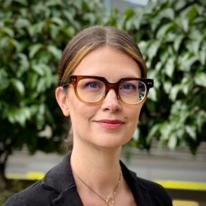 A portrait of Jessi Goss. Jessi has her hair tied back and is wearing stylish thick framed glasses. The background is lovely green trees.