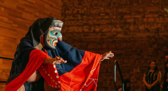 The cover image for the Coast Funds 2024 Annual Report shows a First Nations person dressed in regalia performing at a ceremony. Their regalia is a mix of bright reds, deep blues and black. They are wearing a large mask that is painted with a kaleidoscope of teal, blue, orange, red, and black.
