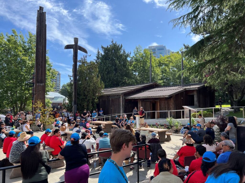 Many people gather outside the Mungo Martin Longhouse. A lot of the people gathering are First Nations Guardians. There is a person, Yuxwelupton Qwal’ qaxala Bradley Dick, who is speaking to the audience. In the background there are three totem poles.