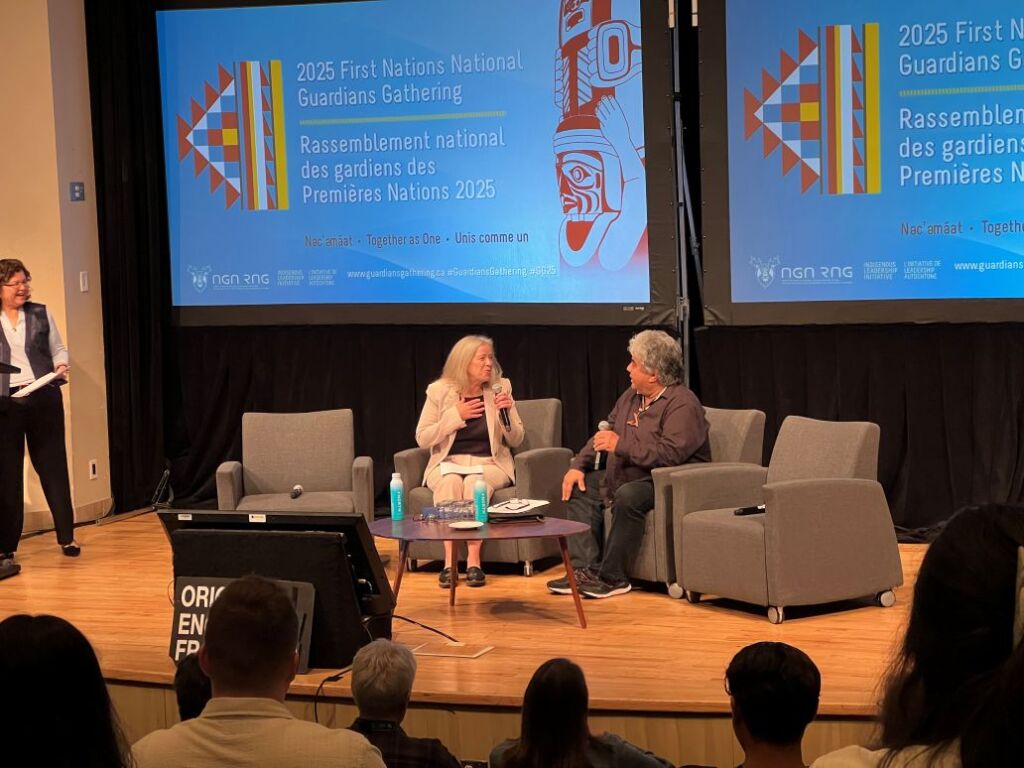 Two people, Hon. Randene Neill M.L.A. and Miles Richardson O.C., sit together on stage in deep discussion.