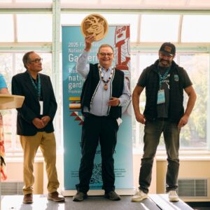 Three people stand together on a small stage. They are Troy Woodhouse, Danny Yakeleya, and Chief Frank Brown. Chief Brown is holding an award. All three people are smiling.