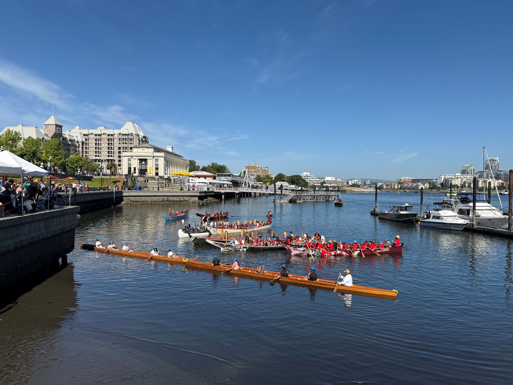 Seven canoes gather together at the corner of a harbour. They are positioned to face a large group of people who are gathered on the land. The people in the canoe are holding paddles carved with beautiful designs. In this photo, you can see that the people in the canoe are speaking to the people on the land.