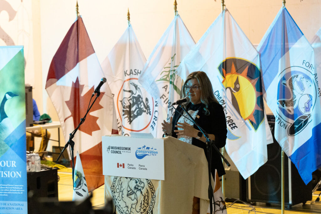 A lovely bright and colourful photo of Mushkegowuk Council Deputy Chief Natasha Martin standing at a podium speaking to a crowd. She is smiling while talking and using her hands. Behind her is a line of many bright flags representing the different Nations of the Omushkego Wahkohtowin PFP. 