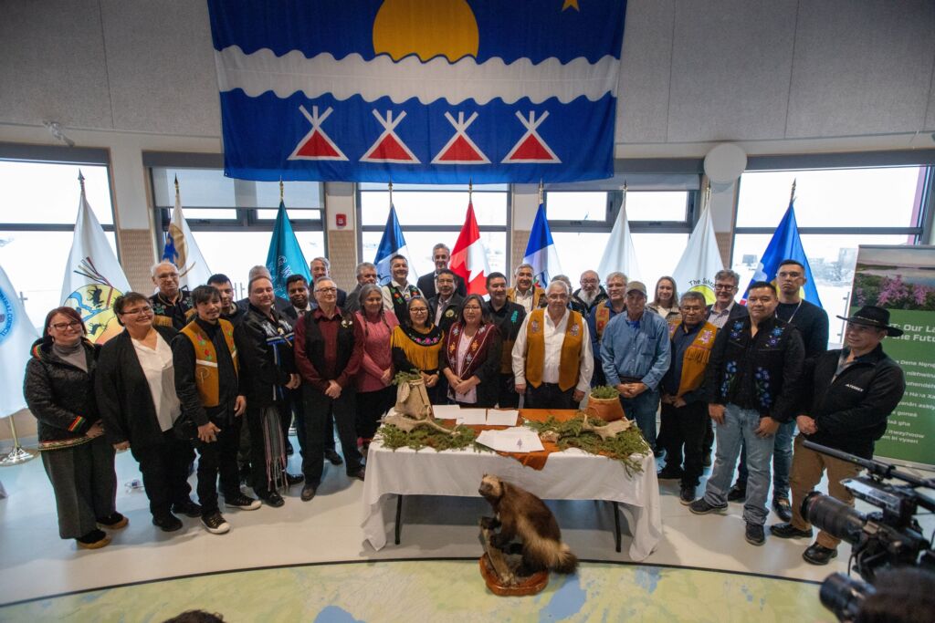 A large group of people stand together surrounding a table with many official documents on it. The group are dressed in a range of regalia, formal business wear, and business casual. Behind the group is a range of different flags representing the different Indigenous Governments of the Northwest Territories. 