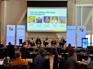 Four representatives of the PFPs in Canada sit at a panel before a large audience. The facilitator, Eddy Adra, is on one side and is directing questions to the panelists.