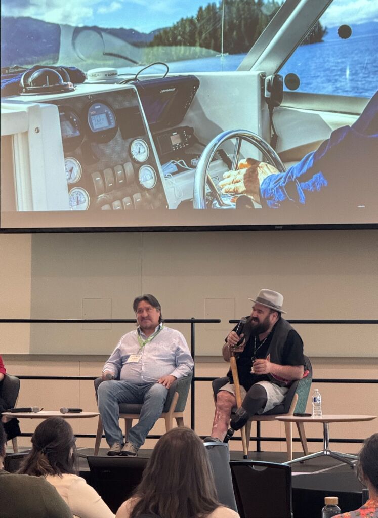 Two people, Steve Nitah and Dallash Smith, sit on stage before a large crowd. Behind them is a projected image of a Guardian driving a boat. 