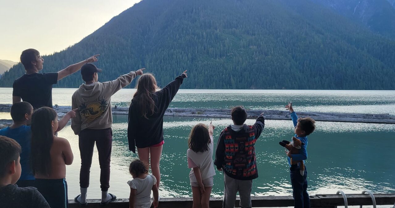 A group of young people gather by a creek and look out at the mountains. Many of the youth are pointing at something in the distance. All people are looking away from the camera.