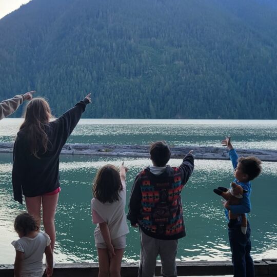 A group of young people gather by a creek and look out at the mountains. Many of the youth are pointing at something in the distance. All people are looking away from the camera.