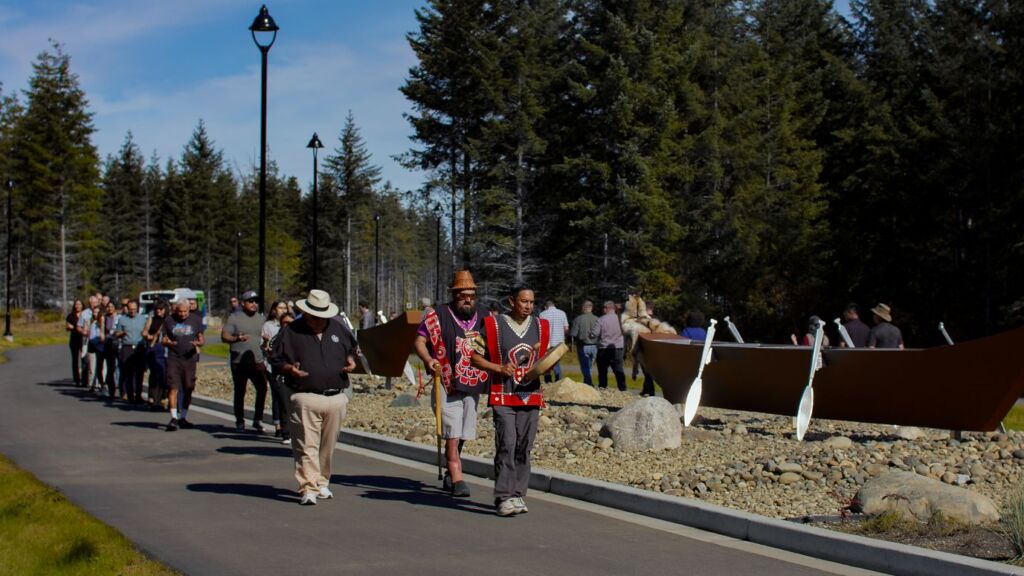 Members of Tlowitsis Nation walking through the newly paved roads of Nenagwas. Beside them are two statues of canoes with paddles. 