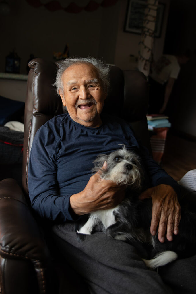 Elder Chief Jacob Nyce and his dog Covid are sitting together on a leather chair. The window beside them is letting in some lovely soft light.