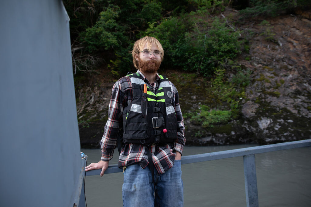 James Griffin is leaning against a rail and standing on a pontoon. He is wearing a flannel shirt and life vest. He is smiling.