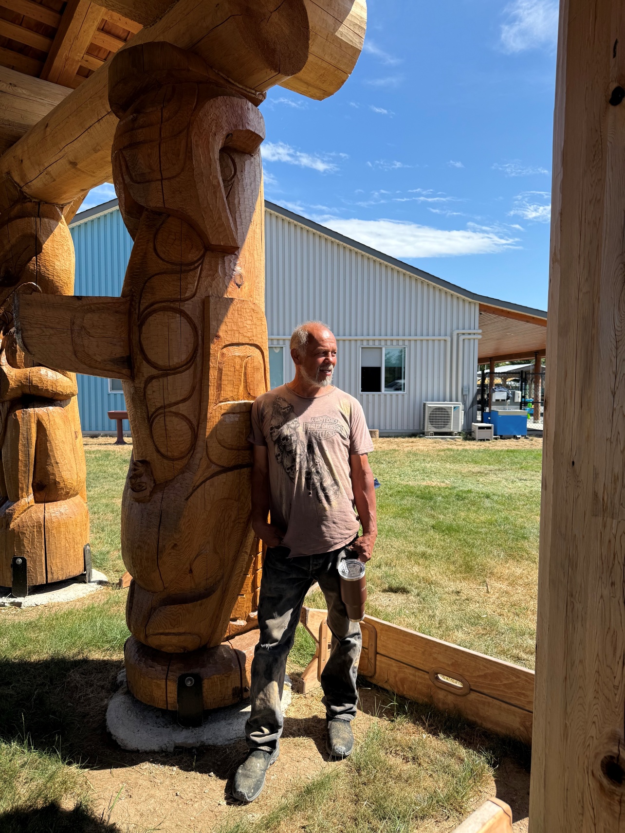 Max stands outside leaning against a pole he carved himself. He is relaxed and looking off in the distance. He holds a coffee travel mug in his right hand.
