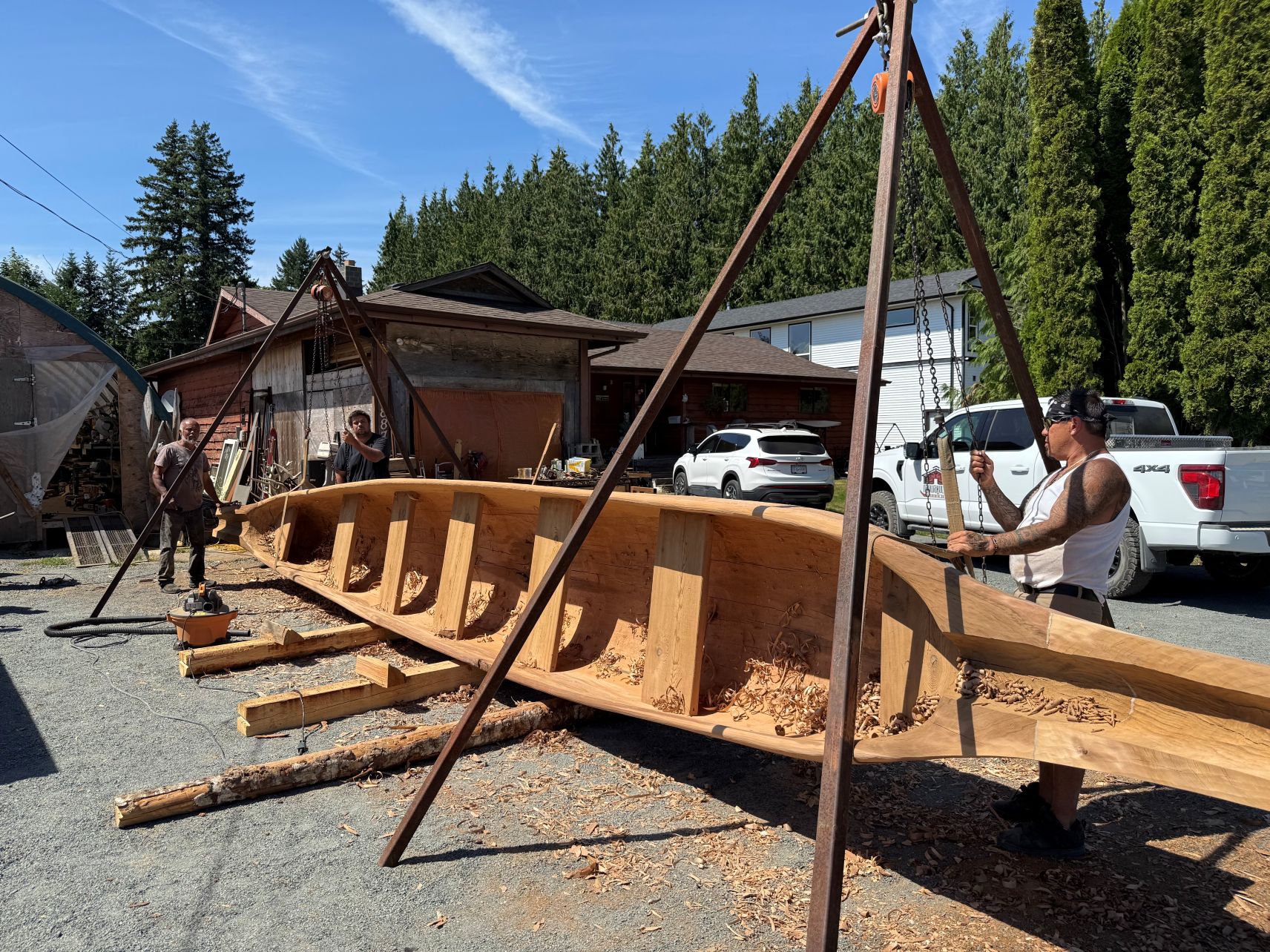Three carvers: Max Chickite, Junior Henderson, and Rayman Shaw are outside Max's home working on a canoe carving. The canoe is connected to a rig and placed on it's side.
