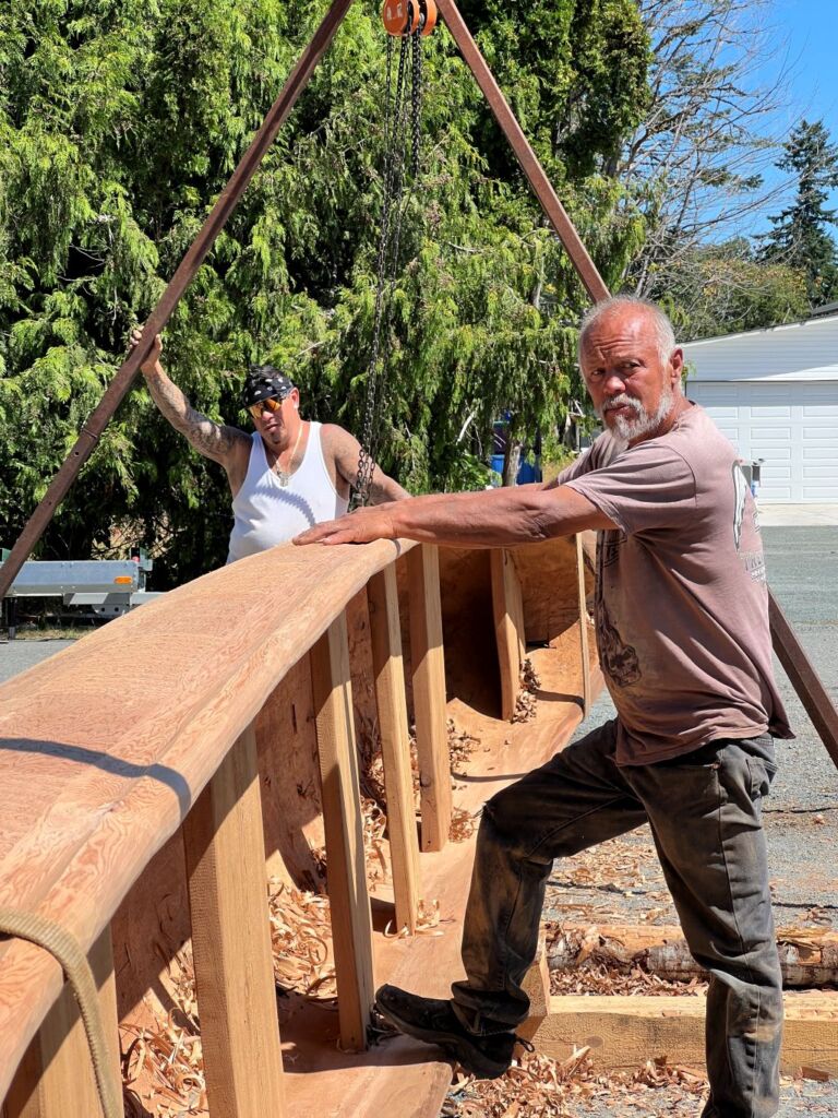 Max stands with his foot resting on the bottom-side of the canoe and both arms balancing on the top-side. He is looking off concentrating on something. Behind him, Junior Henderson has his arm hanging from the A-frame rig used to turn the canoe.