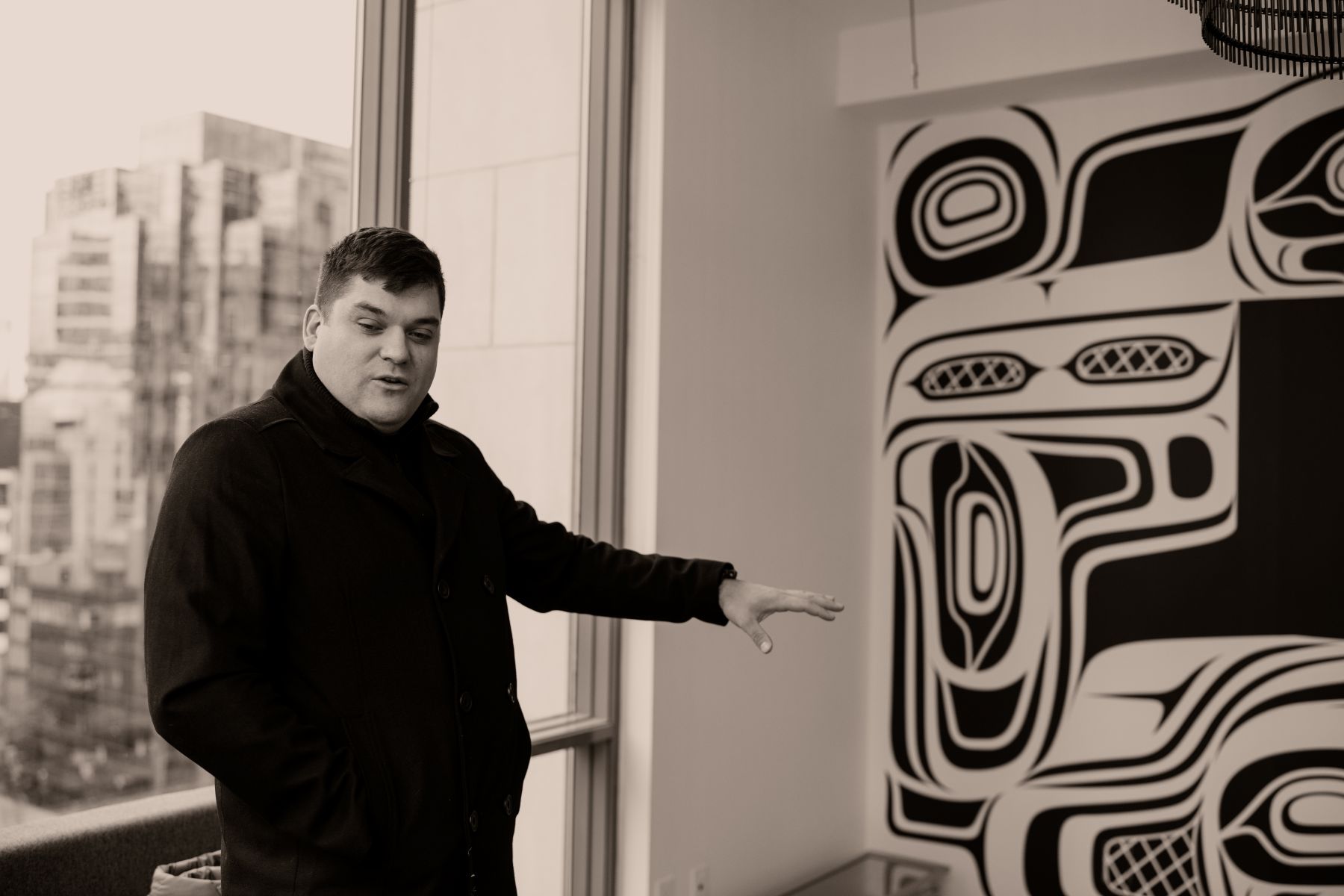 Kolten stands before the mural. He is pointing to one of the figures he designed and explaining the story behind the mural. Kolten is wearing a black coat. The city skyline shines through behind him.