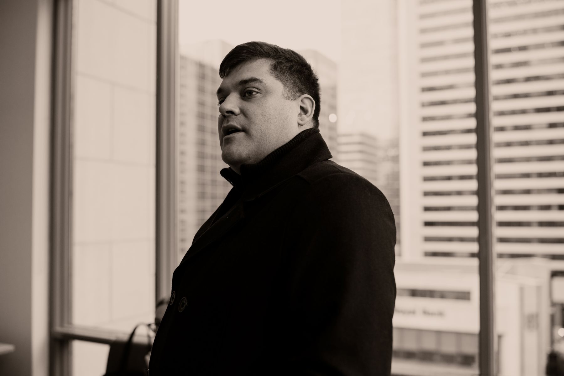 A sepia photograph of Khasalus Kolten Grant. He is wearing a black coat and is standing in front of large windows.