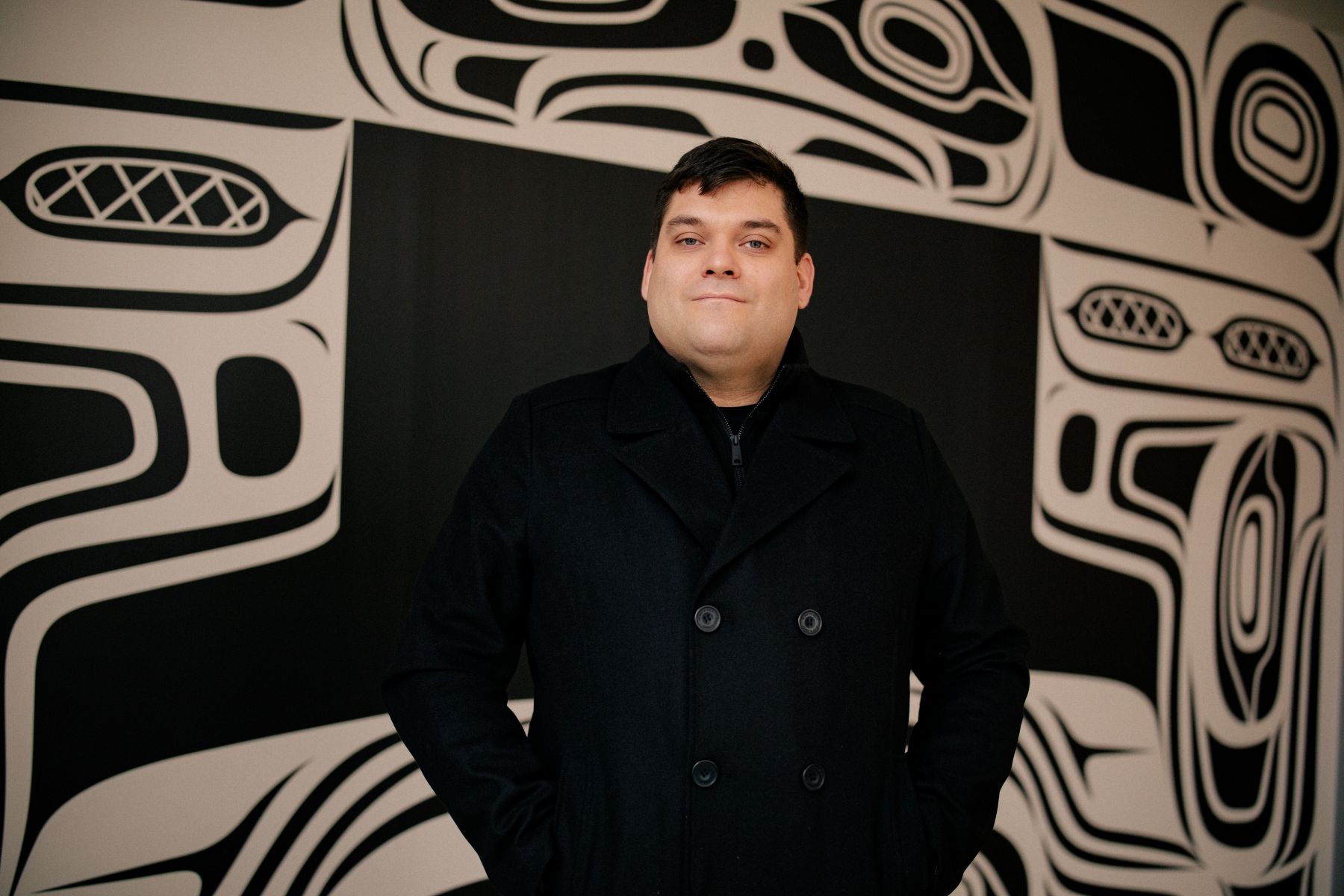 Khasalus Kolten Grant stands before the mural. The mural is black and white, containing four beings and a number of oceanic motifs related to the story of the mural. Each formline is designed in a way to surround a TV at the centre of the wall.