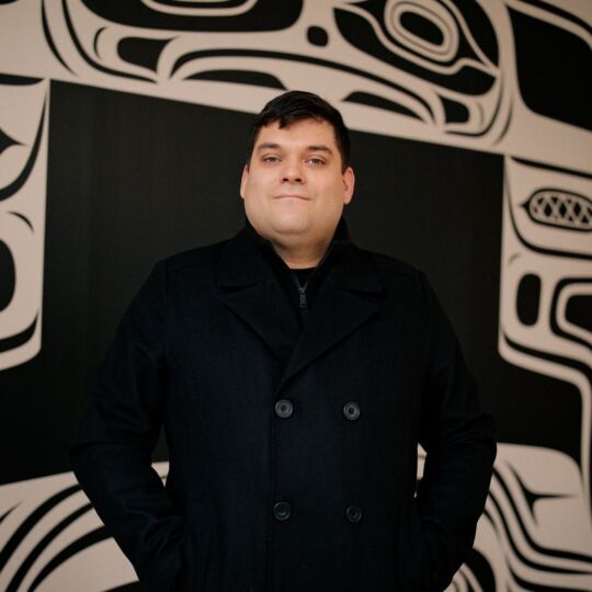 Khasalus Kolten Grant stands before the mural. The mural is black and white, containing four beings and a number of oceanic motifs related to the story of the mural. Each formline is designed in a way to surround a TV at the centre of the wall.