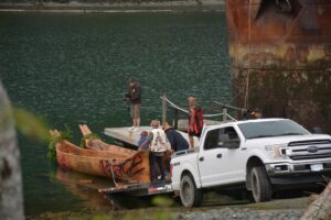 Five people assist with lowering a canoe into the water. The canoe is rigged up on a trailer connected to a utility vehicle.