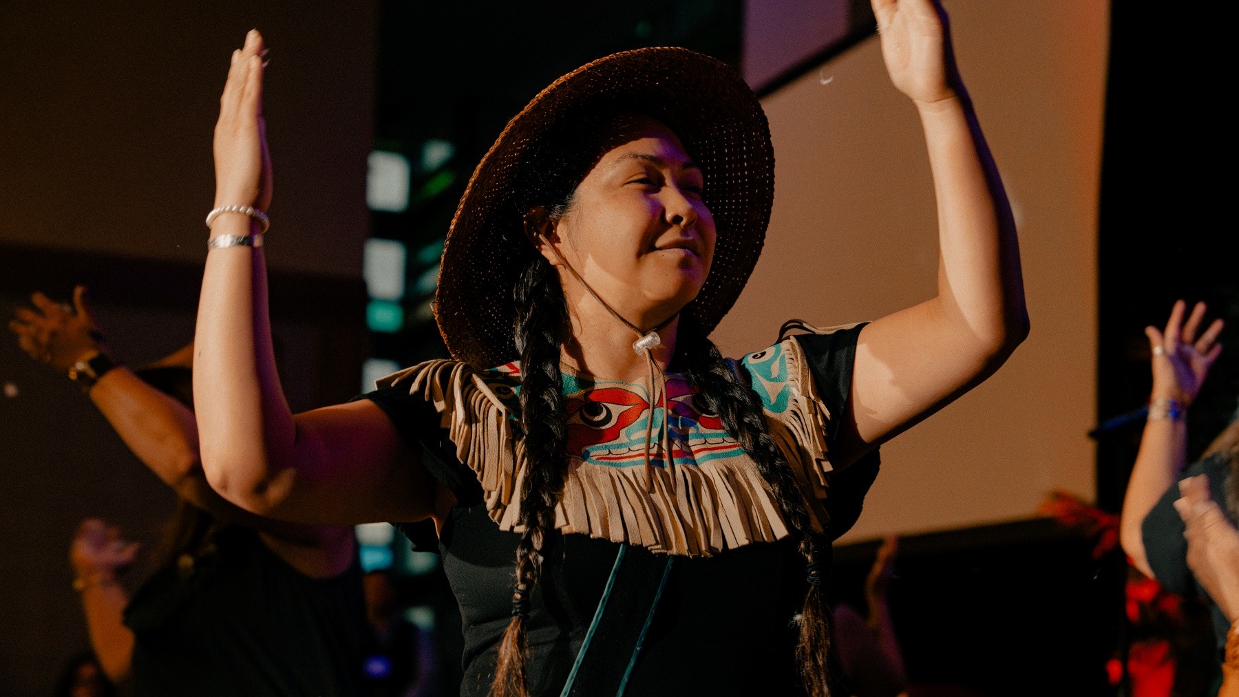 A person, wearing traditional regalia, is dancing. They are smiling and have their arms raised in welcome.