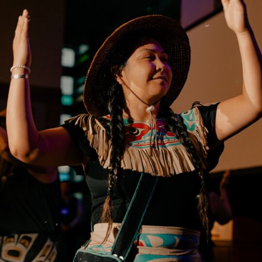 A person, wearing traditional regalia, is dancing. They are smiling and have their arms raised in welcome.