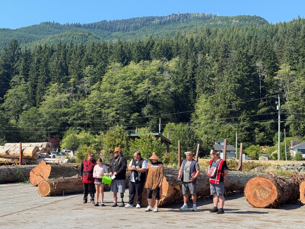 Seven people stand before large cedar logs. Behind them, the forest is green and luscious.