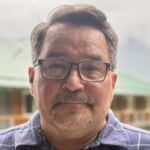 Merv Child smiles at the camera. He is wearing glasses and a button down plaid shirt.