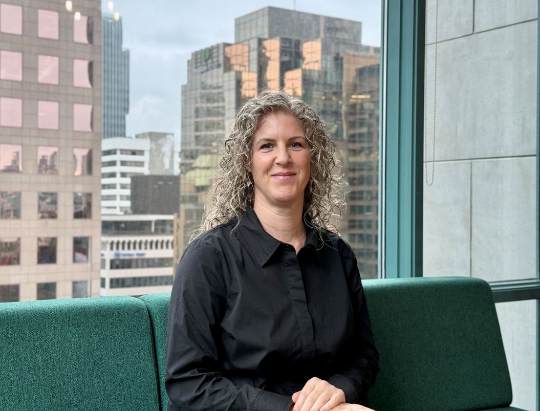 Eau-Vive sits on a green couch and smiles at the camera. She is wearing a black button-down shirt and has lovely curly hair. Behind her is a window that shows off the city skyline of Vancouver.