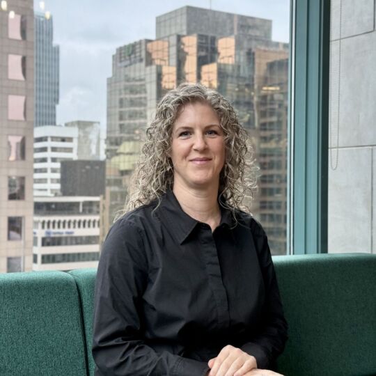 Eau-Vive sits on a green couch and smiles at the camera. She is wearing a black button-down shirt and has lovely curly hair. Behind her is a window that shows off the city skyline of Vancouver.
