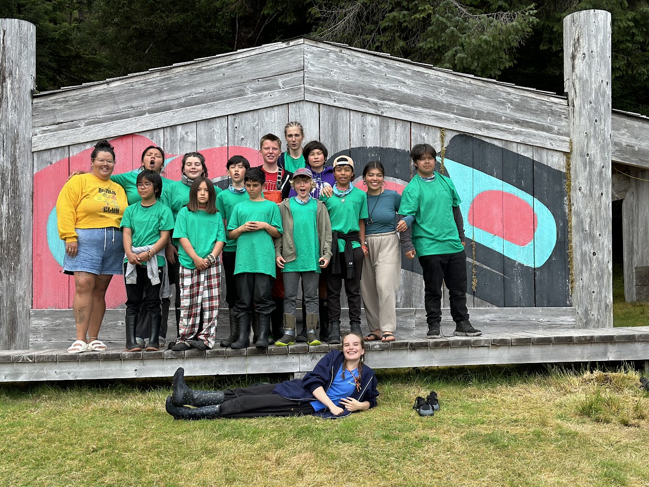 Youth wearing matching green shirts are posing in front of a long house. 