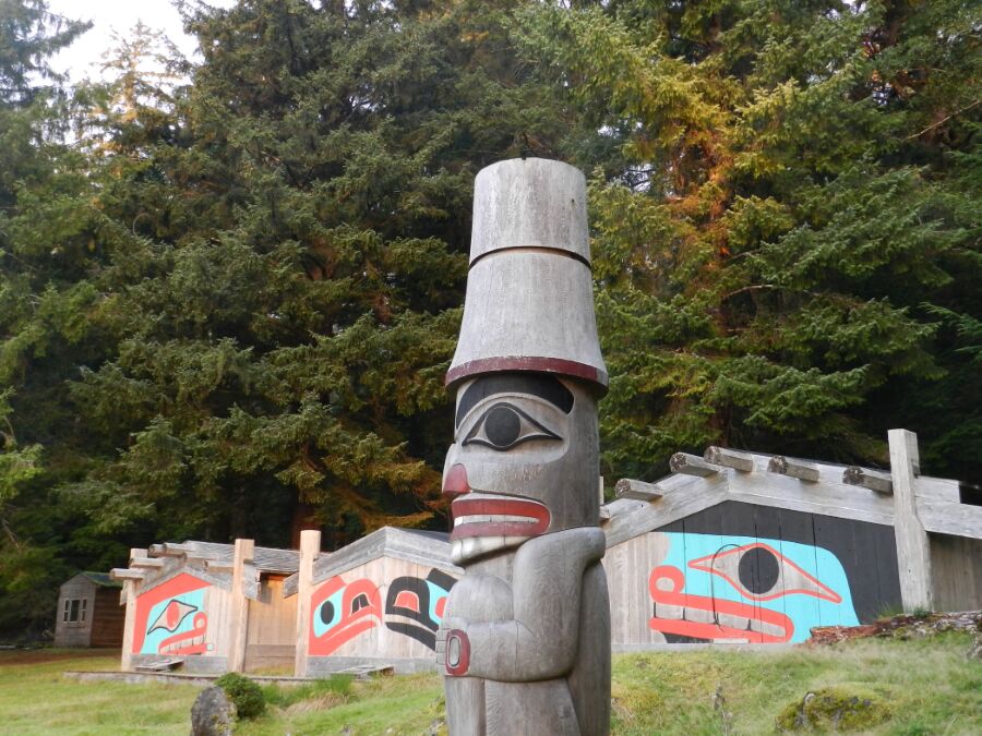 In the foreground is a Haida pole. Behind, three long houses and lots of lovely trees.