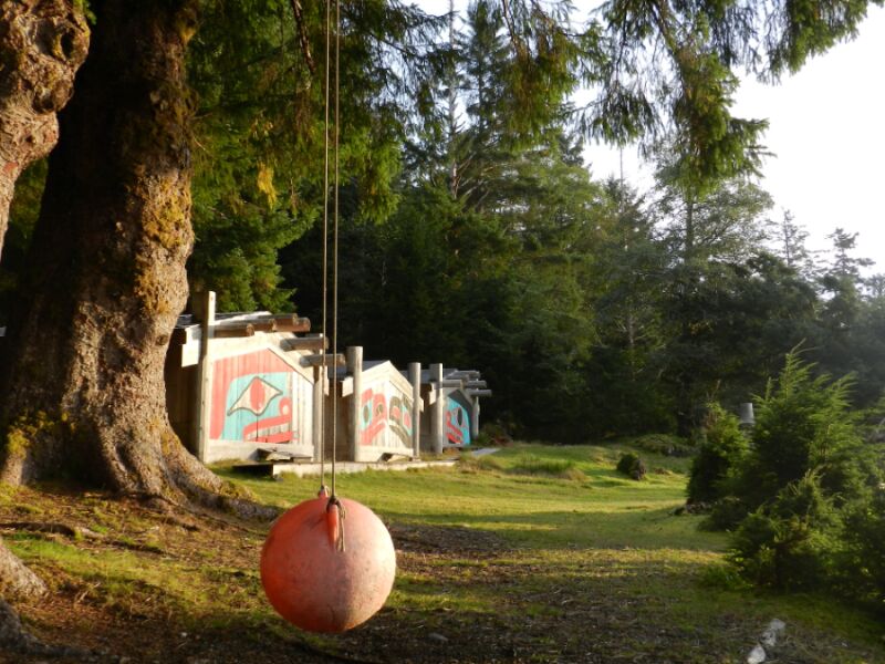 The photograph is outside and depicts a swing in the foreground. Behind, in the background, is arow of long houses with green and red painted ovoid shapes covering the front.