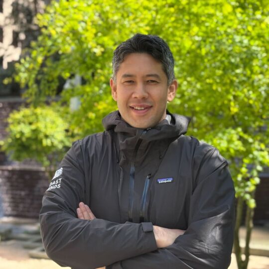 Hide is smiling at the camera. It's sunny outside and there are very bright green trees behind him. Hide has short hair and is wearing an outdoor jacket.