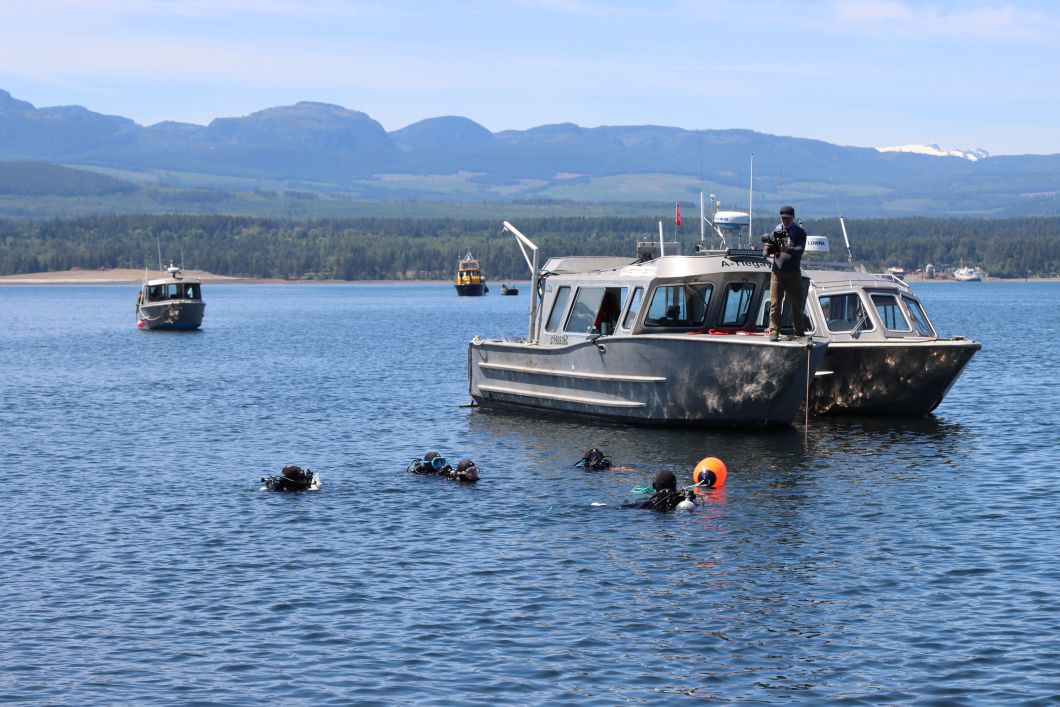 Five divers are floating in the water. Beside them is a boat. Beautiful mountains are in the distance. 