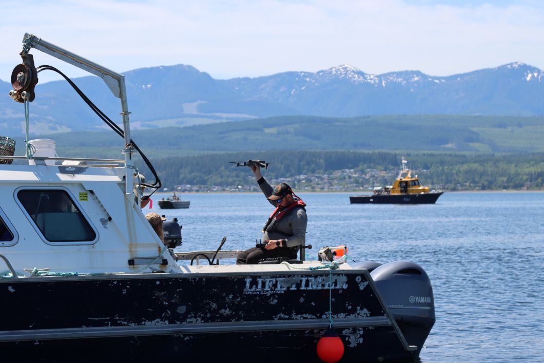 A man is wearing a life jacket and hat while sitting on the back of a boat. He is holding a drone in his right hand and operating it with a a controller in his left hand. 