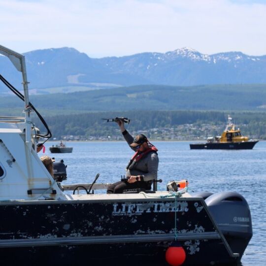 A man is wearing a life jacket and hat while sitting on the back of a boat. He is holding a drone in his right hand and operating it with a a controller in his left hand.
