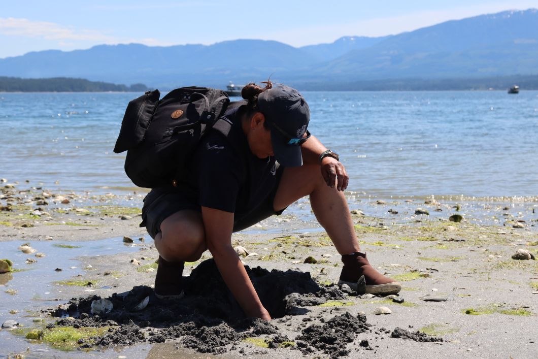 Krissy is crouching down by the sand. She is resting one arm on her leg and then digging into the sand with her other hand. She is wearing a backpack and hat. 