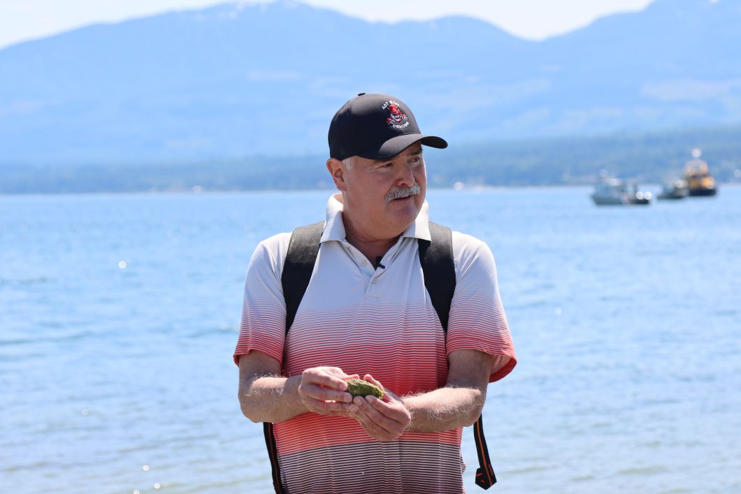 Richard is wearing a hat and a polo shirt. He is holding a clam between his two hands and is talking to a group of people (off camera)